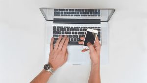 Notebook e mãos no teclado com celular em uma das mãos