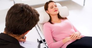 Psicólogo homem de costas sentado escrevendo enquanto paciente está deitada de olhos fechados em estado de hipnose.