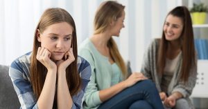 Menina jovem com ansiedade social sem interagir com outras duas meninas sentadas no sofá