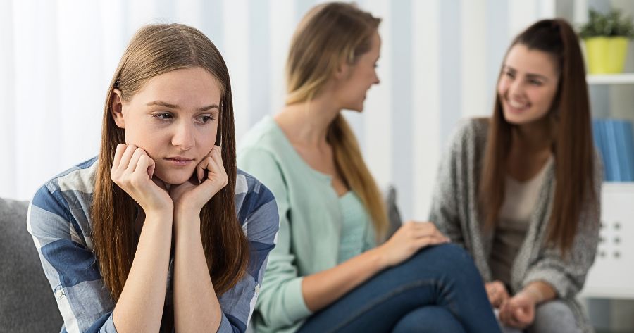 Menina jovem com ansiedade social sem interagir com outras duas meninas sentadas no sofá