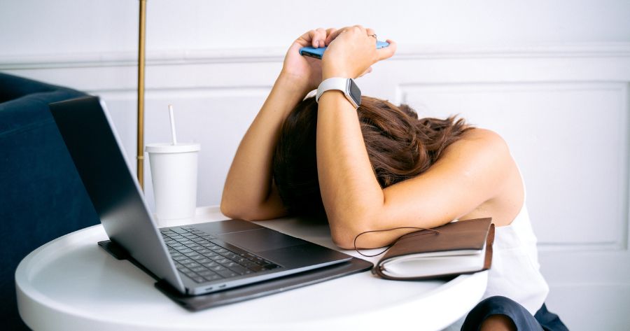 Uma mulher de cabeça baixa com dois cotovelos se apoiando na mesa de frente de um laptop procrastinando o trabalho.