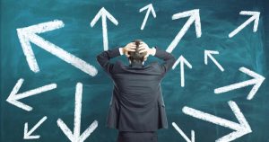 Executivo com as duas mãos na cabeça olhando um quadro com várias setas, remetendo a ideia de dúvida quanto a escolha de uma nova carreira.