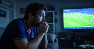 Torcedor assistindo partida de futebol na televisão com expressão de tensão e ansiedade, ambiente escuro, luz da TV refletindo no rosto.